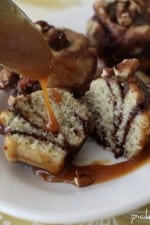 A few caramel turtle sugar brookies with one being cut in half to show the cookie dough and brownie swirl. It is being topped with caramel drizzle from a spoon and there are also chopped pecans on top.