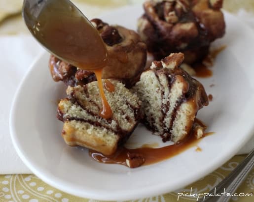 A few caramel turtle sugar brookies with one being cut in half to show the cookie dough and brownie swirl. It is being topped with caramel drizzle from a spoon and there are also chopped pecans on top.