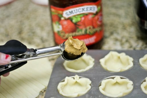 A cookie scoop is used to spoon peanut butter into a tin filled with mini pie crusts.
