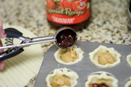 A cookie scoop drops jelly into mini pie crusts filled with peanut butter.