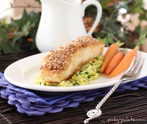 A Chilean sea bass fillet crusted with pecans and Dijon on a plate, served over zucchini slaw and baby carrots.