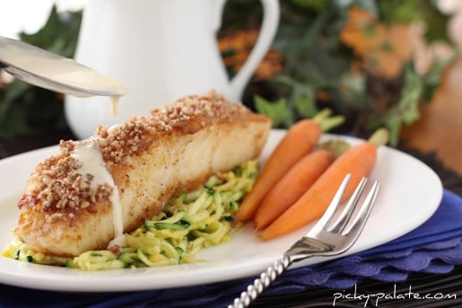 A Chilean sea bass fillet crusted with pecans and Dijon on a plate, served over zucchini slaw and baby carrots.