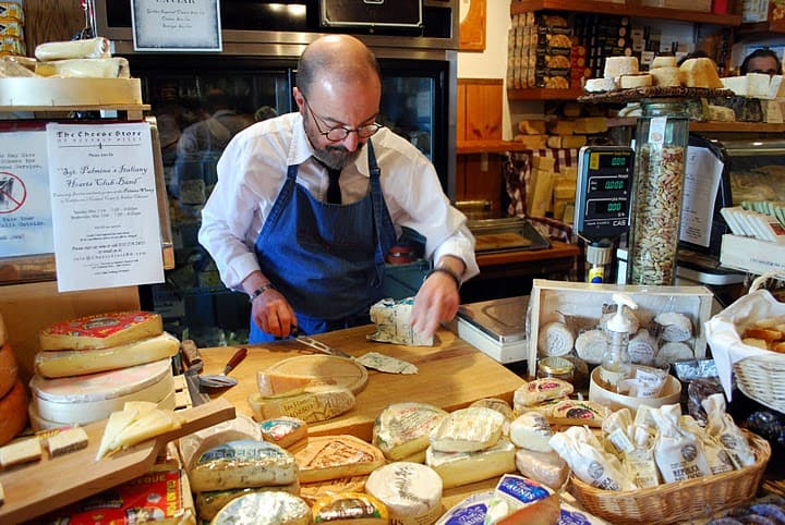 A Man in a Blue Apron Behind the Counter of the Cheese Store