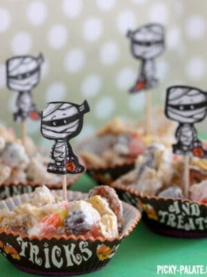 Clusters of mummy munch Halloween Chex mix in small cupcake liners decorated with mummy cutouts on toothpicks.