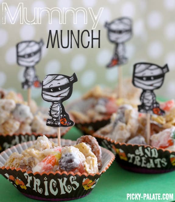 Clusters of mummy munch Halloween Chex mix in small cupcake liners decorated with mummy cutouts on toothpicks.