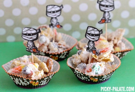 Clusters of mummy munch Halloween Chex mix in small cupcake liners decorated with mummy cutouts on toothpicks.