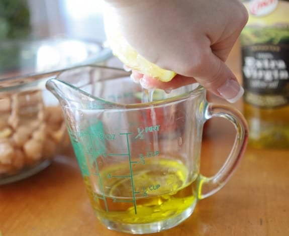 A hand squeezes a lemon over a measuring cup filled with olive oil.