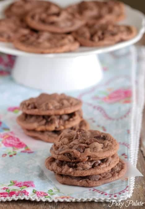 Chocolate malted chip cookies stacked on parchment paper, with more cookies on a cake stand in the background.