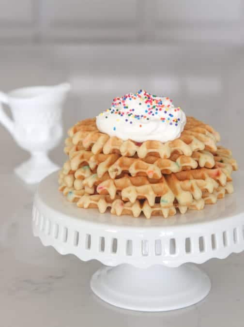 A Pile of Birthday Cake Waffles on a Cake Stand in Front of a Container of Syrup