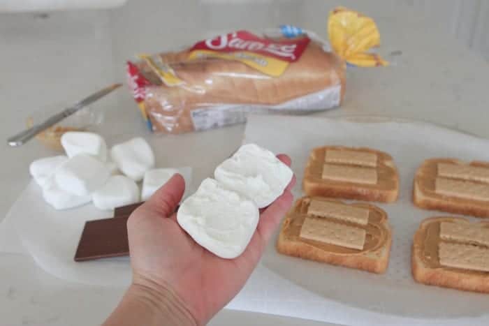 A hand holding up two marshmallows with bread slices topped with peanut butter and Graham crackers in the background.