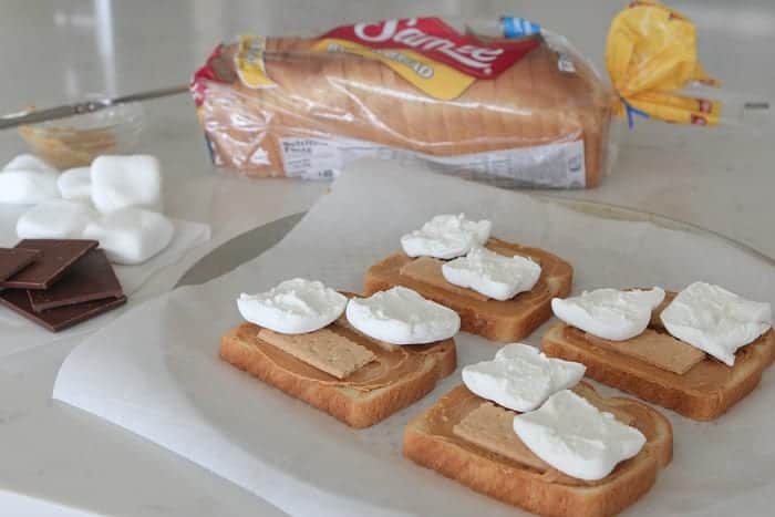 Slices of bread topped with peanut butter, Graham crackers, and marshmallows on a parchment-lined dish.
