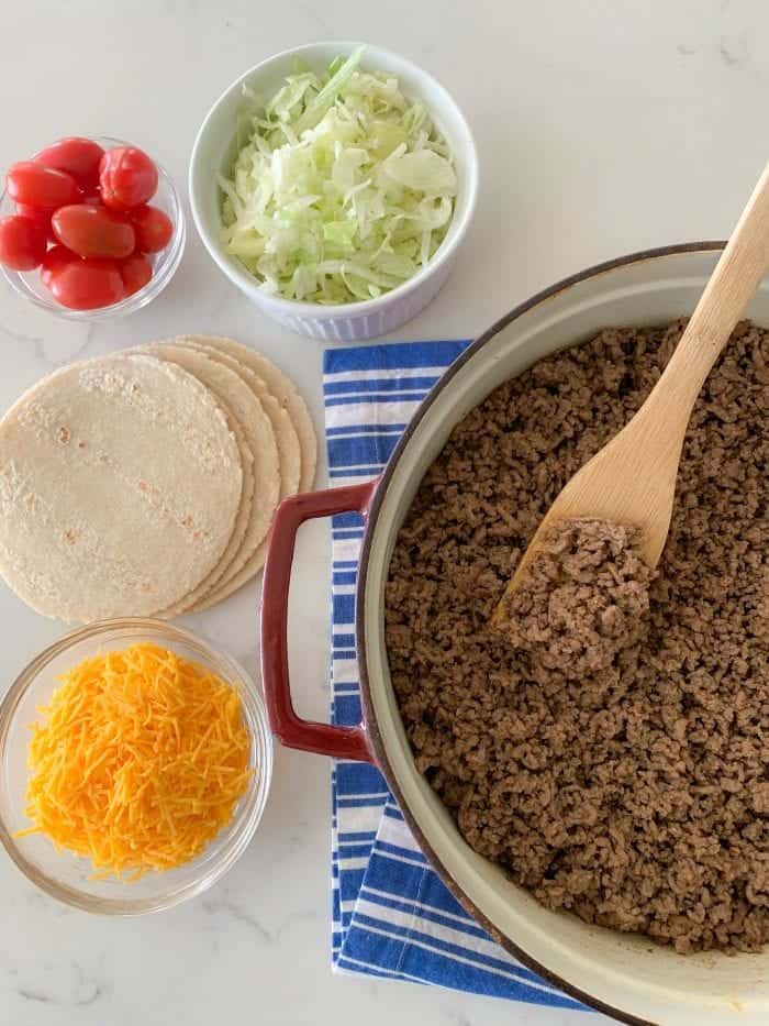 a pot with taco meat next to tomatoes, salad, tortillas and cheese