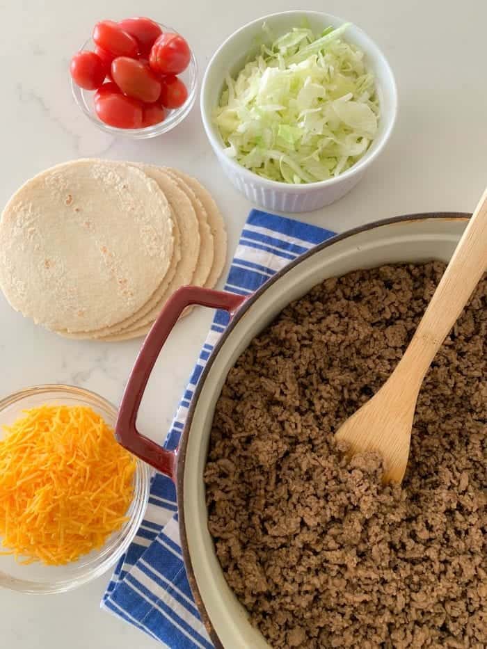 a pot with taco meat next to tomatoes, salad, tortillas and cheese
