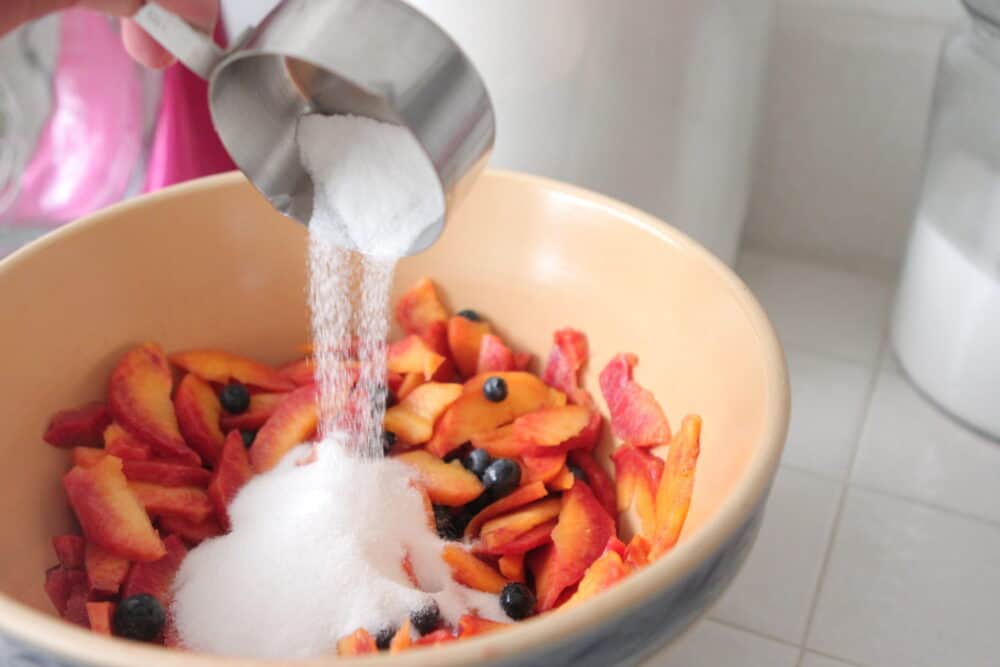 adding sugar to sliced peaches and blueberries in mixing bowl